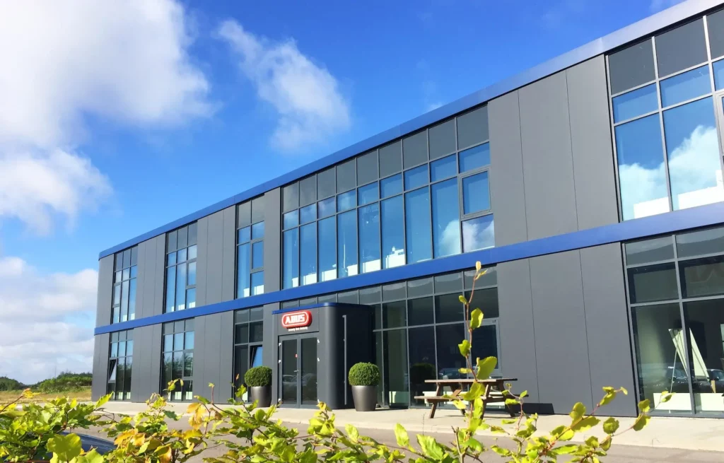 The image shows a modern office building with large glass windows and a dark facade, accented with blue details. In front of the entrance are two large pots of plants and a picnic table, surrounded by green bushes under a clear blue sky.
