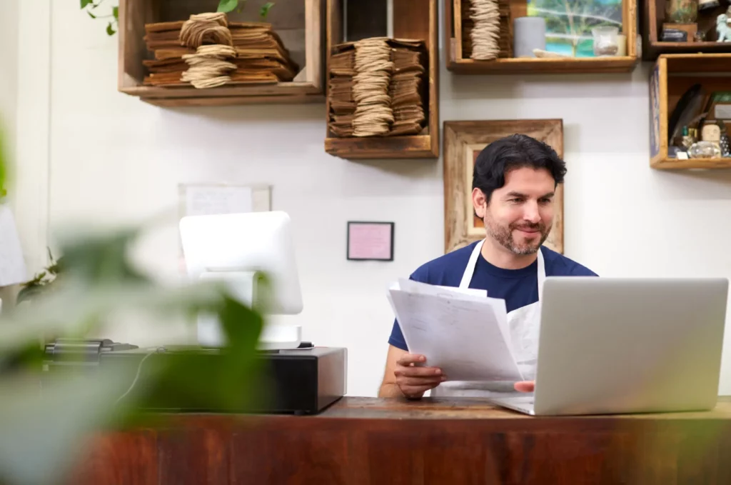 Male shopkeeper with white apron going through paperworks in front of laptop in a shop setting.