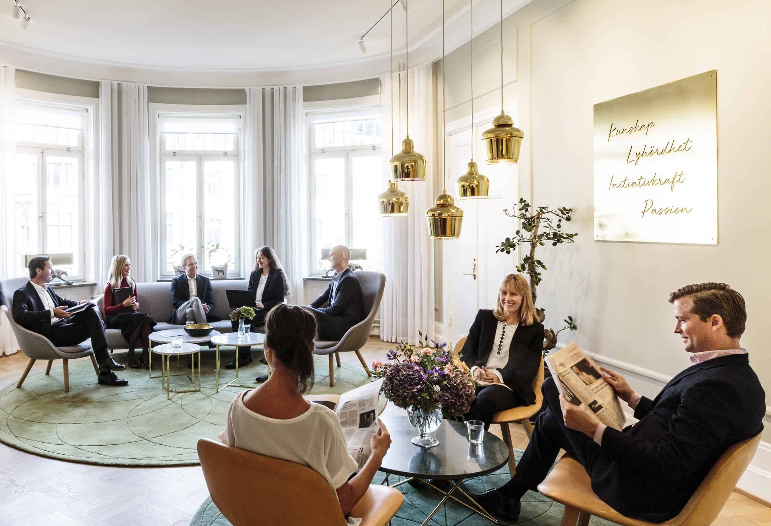 Neatly dressed men and women sit around two tables. One man is reading a newspaper.